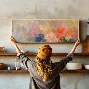 May include: A rectangular abstract painting in a light wood frame featuring thick, textured brushstrokes in shades of muted gray, soft pink, vibrant orange, and deep blue, displayed horizontally against a neutral wall above wooden shelves.