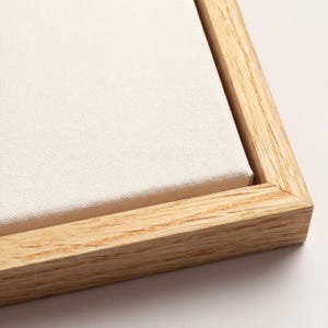 May include: Close-up of a blank canvas stretched over a wooden frame. The canvas is a light cream color, and the frame is made of light-colored wood. The corner of the canvas and frame are visible, showing the construction.