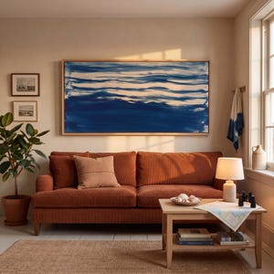 May include: A large rectangular abstract painting with blue and white horizontal brushstrokes hangs above a burnt orange sofa. A wooden coffee table holds a lamp, a bowl of seashells, binoculars, and books. A potted plant stands to the left.