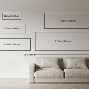 a wall with a series of framed artworks or photographs of varying sizes, ranging from 24 inches to 72 inches in width. In front of the wall, there is a beige couch or sofa, providing a contrast to the wall display.