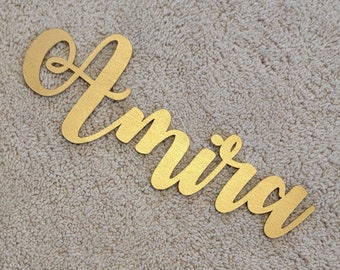 May include: Gold-colored wooden name sign with the name "Amirah" written in cursive script.