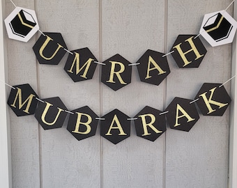 May include: Black and gold banner with the words "Umrah Mubarak" in gold letters on black hexagonal shapes. The banner is strung with white string and has two white hexagonal shapes with a black and white image of the Kaaba on each end.