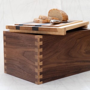 Può includere: Un portapane in legno con giunti a coda di rondine, sormontato da un tagliere in legno. La tavola ha un design a strisce nere e contiene pane a fette e un coltello piccolo. La scatola e la tavola sono in legno chiaro e scuro.