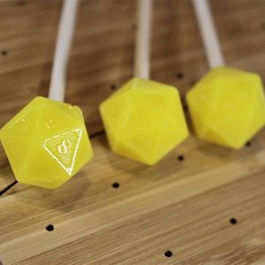 May include: Three yellow, candy-like dice on sticks. The dice are shaped like a twenty-sided die, also known as a d20.