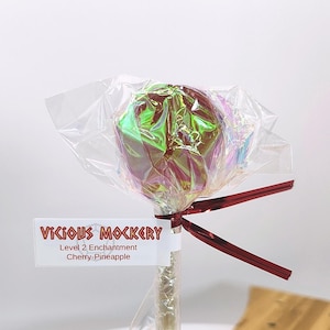 Può includere: Un lecca lecca con un involucro olografico e un nastro rosso. L'etichetta recita "Vicious Mockery, Level 2 Enchantment, Cherry-Pineapple". La caramella è su un bastoncino trasparente.