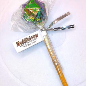 May include: A root beer float lollipop with a holographic wrapper and a wooden stick. The label reads "Muddlebrew Level 2 Transmutation Root Beer Float". Silver ribbons are tied to the stick.