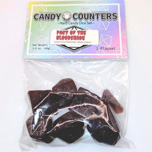 Op de afbeelding: Een doorzichtige plastic zak met donkerbruine, onregelmatig gevormde harde snoepdobbelstenen. De zak heeft een kleurrijke koptekst met de tekst "CANDY COUNTERS" en "PACT OF THE BLOODSHADE". Het nettogewicht is 40g.