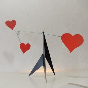 May include: A black metal stand with three legs holds a mobile with three red paper hearts. The hearts are suspended from a thin wire.