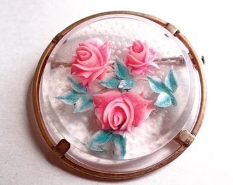 May include: A vintage brooch with a clear, circular design. It features a cluster of pink roses and blue-green leaves, set against a textured background, and framed by a gold-toned metal border. A classic piece of jewellery.