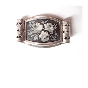 Peut inclure: Un bracelet en métal argenté avec un charme rectangulaire en émail noir orné d'un motif floral blanc. Le charme a un motif floral blanc sur fond noir.