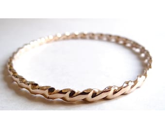 Puede incluir: Un brazalete dorado con un diseño de cuerda trenzada. El brazalete es circular y parece estar hecho de metal. El patrón trenzado le da al brazalete una apariencia texturizada. Esta joya es adecuada para el uso diario.