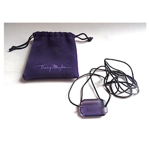 Könnte beinhalten: Eine Halskette mit einem violetten Kristall-Anhänger, einer schwarzen Schnur und einem violetten Samtbeutel mit dem aufgedruckten Text "Thierry Mugler".