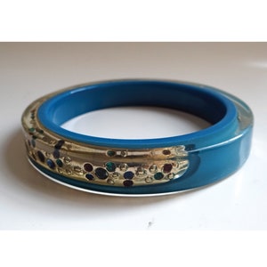 Pu&ograve; includere: Un braccialetto blu turchese con una fascia dorata e punti colorati. Il braccialetto &egrave; in plastica e ha una finitura lucida.