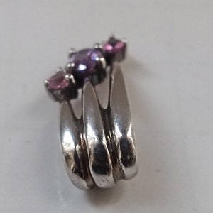 Pode incluir: Um pingente de prata com tr&ecirc;s pedras preciosas roxas e cor-de-rosa engastadas em um design curvo.