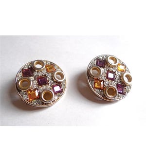 May include: A pair of circular earrings with a silver finish. Each earring has a central pattern of square and round gemstones in purple, orange, and clear, encircled by smaller clear stones.