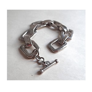 Può includere: Bracciale color argento con una serie di maglie rettangolari. Il bracciale ha una chiusura a toggle. Il metallo ha un aspetto strutturato, che gli conferisce un aspetto vintage o artigianale. Il bracciale è esposto su una superficie chiara.