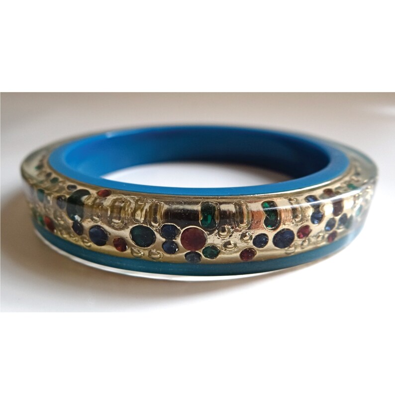 Pu&ograve; includere: Un braccialetto turchese con un designo in oro e resina trasparente. Il design presenta una fascia d'oro con un motivo di piccoli strass colorati.