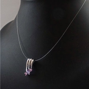 Pode incluir: Um colar de prata com um pingente que apresenta tr&ecirc;s pequenas pedras preciosas de cor p&uacute;rpura. O colar est&aacute; em uma corda invis&iacute;vel transparente.