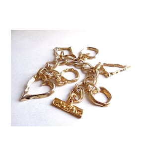 May include: A gold-toned charm bracelet featuring various shapes, including a heart, rings, and a toggle clasp. The bracelet is made of linked chains and has a textured, hammered appearance. The bracelet is displayed on a white surface.