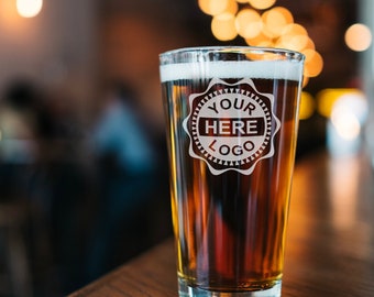 May include: A clear glass pint glass with a frosted design that says "Your Here Logo" filled with a dark amber beer. The glass is sitting on a wooden table.