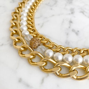May include: A gold chain necklace with multiple layers of chunky chains and white pearls. The necklace features a small gold and clear crystal accent.