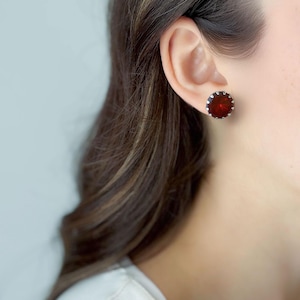 Könnte beinhalten: Nahaufnahme eines Ohrs mit einem runden, dunkelroten Ohrstecker mit silberner Fassung. Der Ohrring hat ein facettiertes Design, das das Licht reflektiert. Die Person hat lange, braune Haare.
