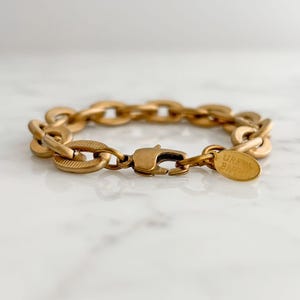 Könnte beinhalten: Goldfarbenes Kettenarmband mit ovalen Gliedern und einem Karabinerverschluss. Das Armband hat eine strukturierte Oberfläche und ein kleines Etikett mit der Aufschrift "GREEN BIRD". Der Schmuck wird auf einer weißen Oberfläche präsentiert.
