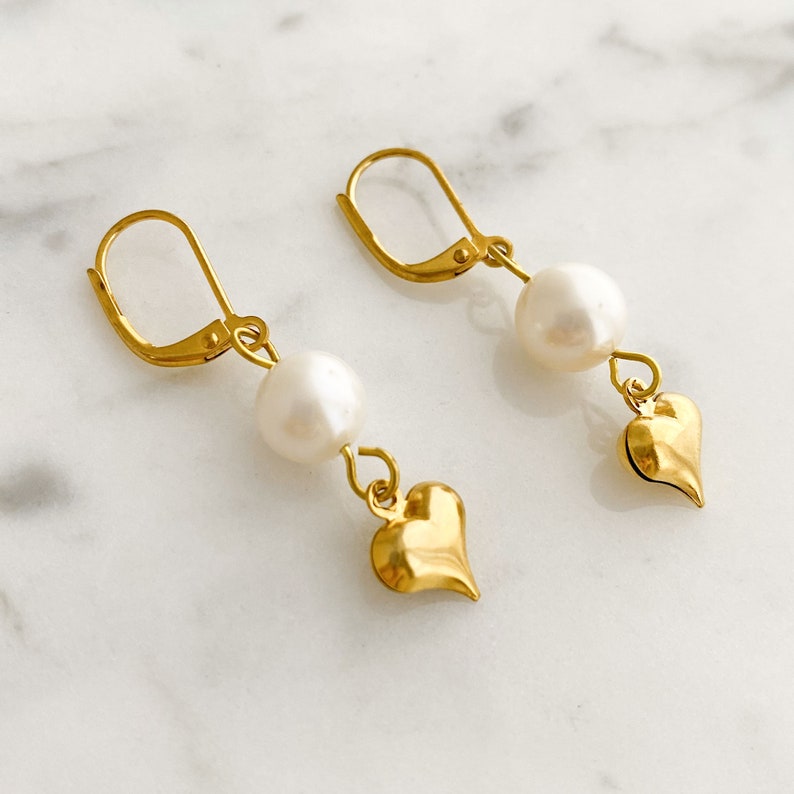 Pode incluir: Brincos de ouro com uma p&eacute;rola e um pingente em forma de cora&ccedil;&atilde;o.