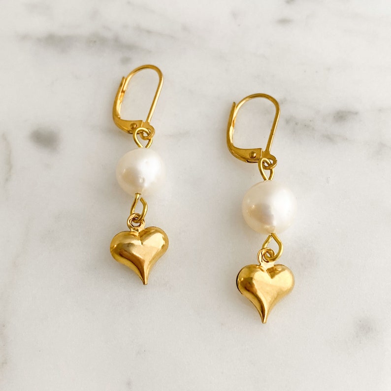 Pode incluir: Brincos de ouro com uma p&eacute;rola e um pingente em forma de cora&ccedil;&atilde;o. Os brincos t&ecirc;m um design simples e elegante.