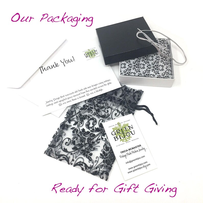 May include: A white card with the text "Thank You!" and a green logo that says "Green Bijou". The card is sitting on top of a black and white patterned gift bag. The gift bag is sitting on top of a white box with a black lid. The box is open and the inside is lined with white tissue paper. The box is sitting on a white surface. The text "Ready for Gift Giving" is written in black letters on the white surface.