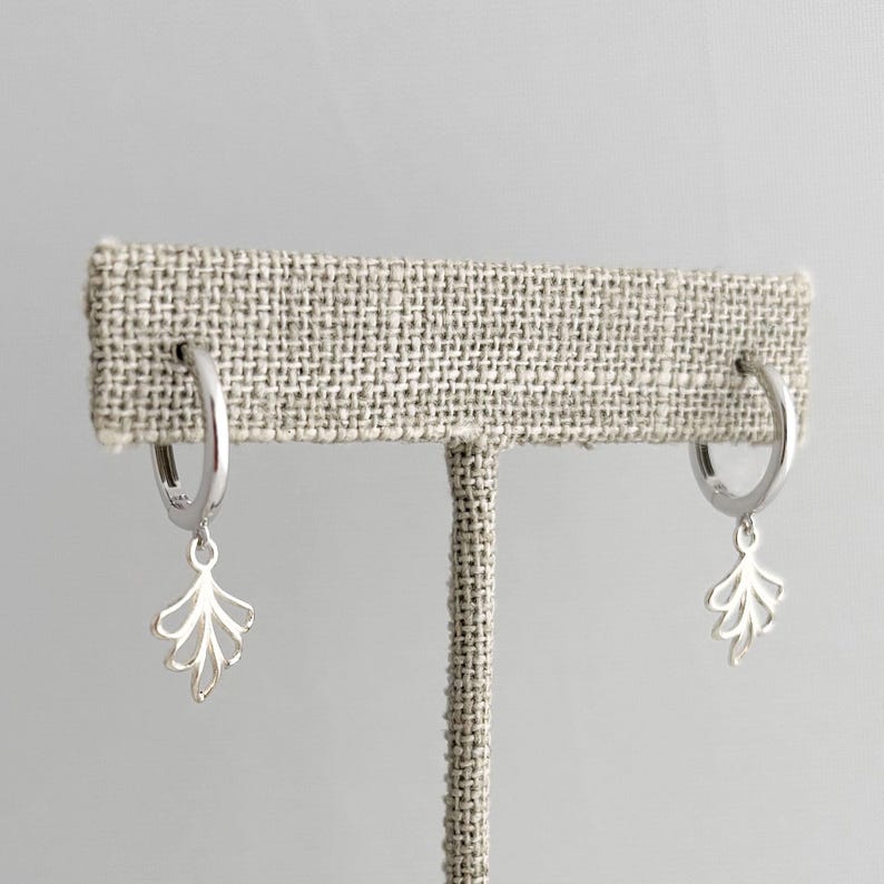 May include: A pair of silver hoop earrings with a delicate leaf charm dangling from each hoop.