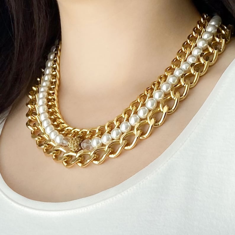 May include: A gold chain necklace with a row of white pearls and a crystal accent. The necklace is layered with two thick gold chains.