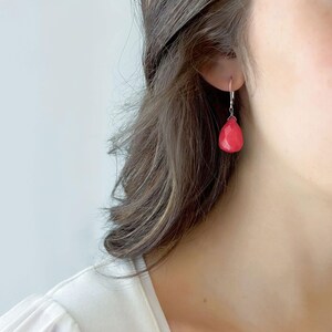 May include: A close-up of a single earring. The earring features a faceted, teardrop-shaped, red gemstone suspended from a silver hook. The gemstone is a vibrant shade of red, catching the light.
