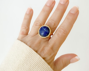 May include: A gold-toned ring featuring a large, circular, deep blue lapis lazuli gemstone set in a smooth bezel. The band of the ring is split into two thin, parallel gold-toned metal wires that connect to the circular setting, creating a simple design.