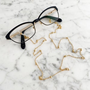 以下が含まれることがあります： 黒とゴールドの縁の眼鏡と繊細なゴールドのチェーンの眼鏡ホルダー。チェーンには、小さなゴールドのビーズが長さに沿って散りばめられています。眼鏡は白とグレーの大理石の表面に置かれています。