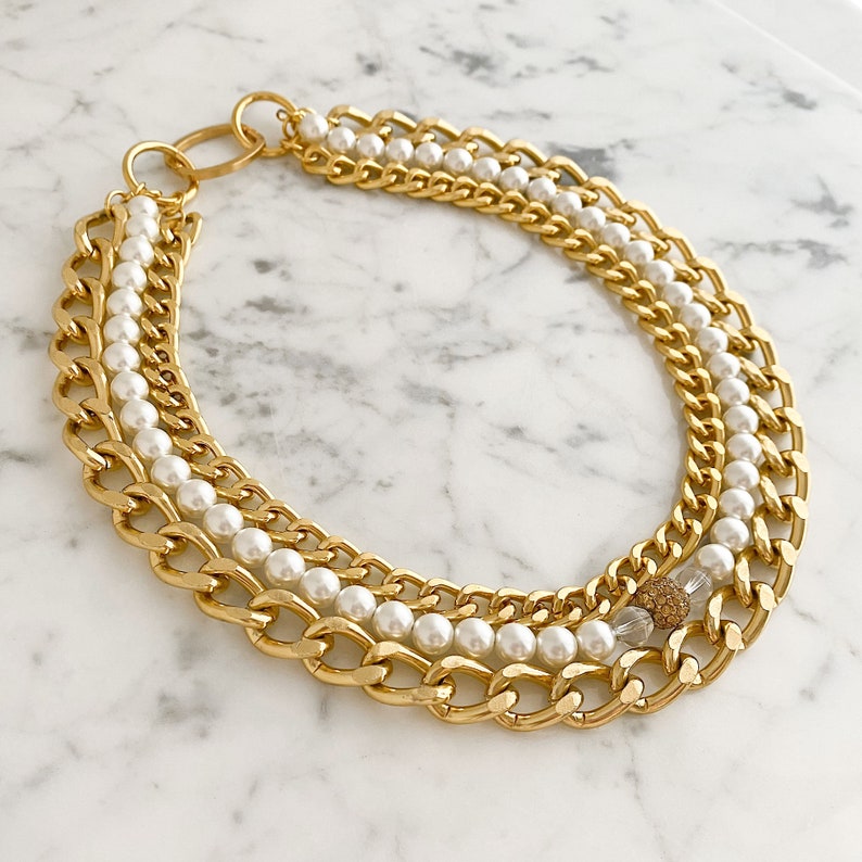 May include: A gold chain necklace with multiple layers of chunky chains and white pearls. A clear crystal with gold glitter accents hangs from the bottom of the necklace.