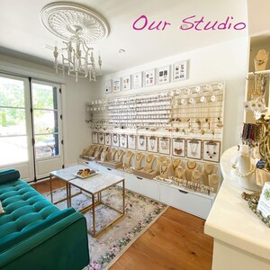 May include: A white wall with the text "Our Studio" in pink lettering. The wall is decorated with jewelry displayed on a pegboard and shelves. The jewelry includes necklaces, earrings, and bracelets. There is a green velvet sofa and a white marble table in the foreground.