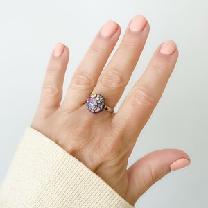 May include: A silver ring featuring a round, multi-coloured stone set in the centre. The stone has a purple, pink, and yellow colour scheme with a glittery finish.