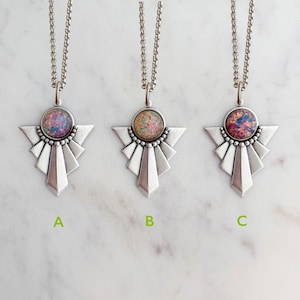 May include: Three silver-toned necklaces featuring geometric fan-shaped pendants with circular cabochon centers. Pendant A has a purple and blue stone, B has an orange and gold stone, and C has a deep red and blue stone. Each hangs on a silver chain.