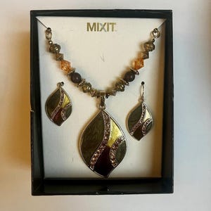 Könnte beinhalten: Ein Schmuckset mit Halskette und passenden Ohrringen, präsentiert in einer schwarzen Box. Die Halskette und die Ohrringe haben ein blattartiges Design mit olivgrüner Emaille, goldenen Akzenten und funkelnden Strasssteinen. Der Markenname "MIXIT" ist sichtbar.