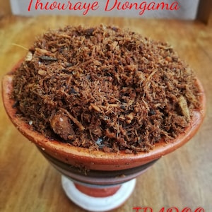 May include: A close-up of a brown ceramic bowl filled with shredded, dried, brown tobacco. The bowl is sitting on a wooden surface. The text "Thiouraye Diongama" is written in red cursive above the bowl, and the text "IBADOO" is written in red cursive below the bowl.