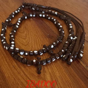 Peut inclure: Chapelet en bois brun foncé avec des points blancs. Les perles sont disposées en cercle, avec un pendentif en forme de croix. Le mot "IBADOO" est visible en texte rouge.