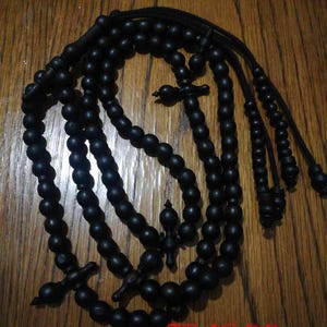 Peut inclure: Chapelet noir avec un pendentif en forme de croix et le mot "TBADOO" en rouge. Les perles sont faites de perles rondes et sombres enfilées avec une ficelle noire. Le fond est une surface en bois.