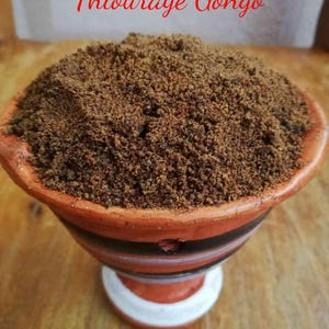 May include: A terracotta bowl filled with dark brown, granular material, possibly a spice or herb blend. The bowl has a striped design and sits on a wooden surface. The text "Thiouraye Gongo" and "IBADOO" are written in red.