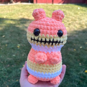 May include: A crocheted plush toy in the shape of a monster with a stitched-on mouth. The toy is pink, yellow, and white with black eyes.