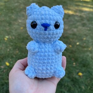 May include: A small, light blue crocheted stuffed animal shaped like a bear. The bear has black eyes, a blue nose, and a fluffy texture.