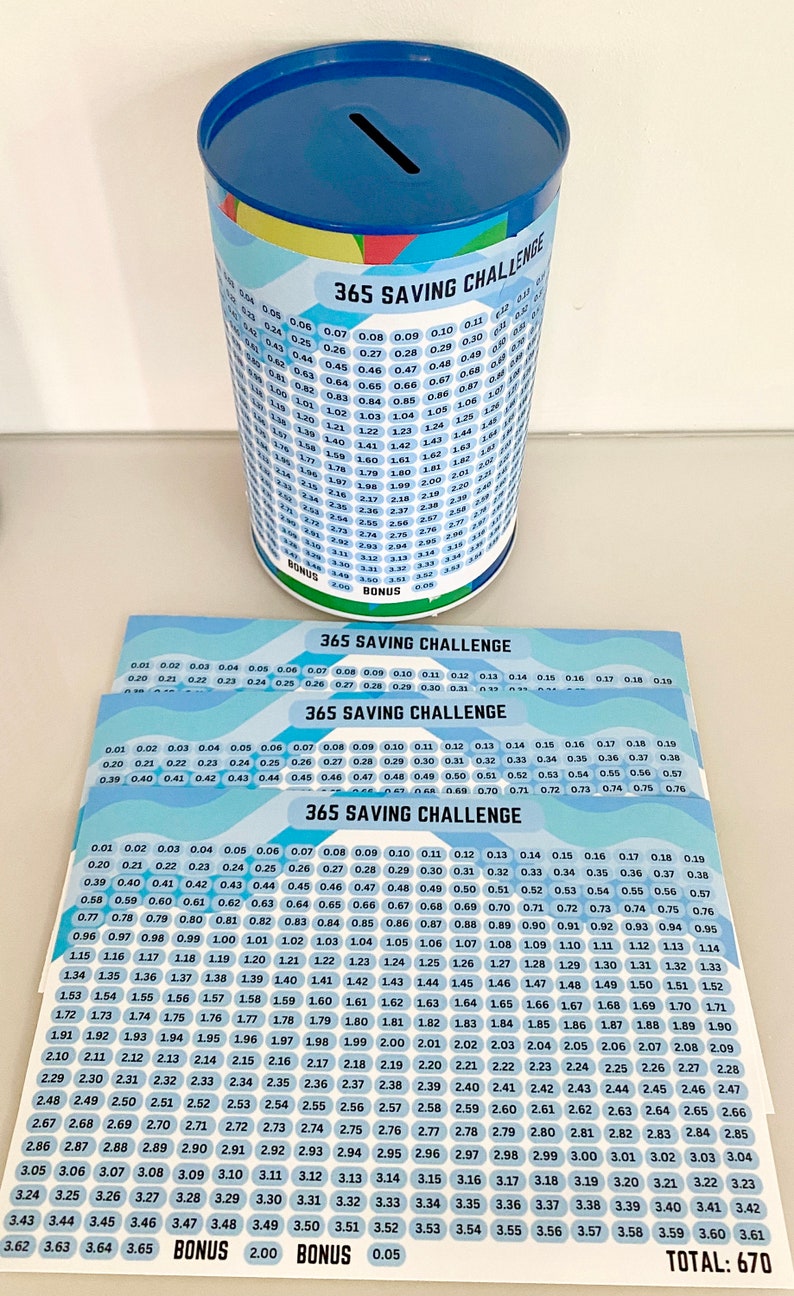 K&ouml;nnte beinhalten: Eine blaue und wei&szlig;e zylindrische Spardose mit einem Schlitz oben. Die Spardose hat eine Tabelle auf der Seite mit dem Text "365 Saving Challenge" und Zahlen von 0,01 bis 3,65. Es gibt drei weitere Bl&auml;tter Papier mit der gleichen Tabelle und dem gleichen Text.