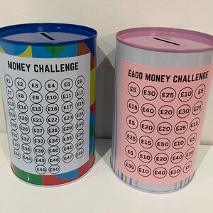 May include: Two metal money challenge piggy banks. The blue and green piggy bank has a money challenge label and a grid of numbers with pound signs. The pink and white piggy bank has a &pound;600 money challenge label and a grid of numbers with pound signs.