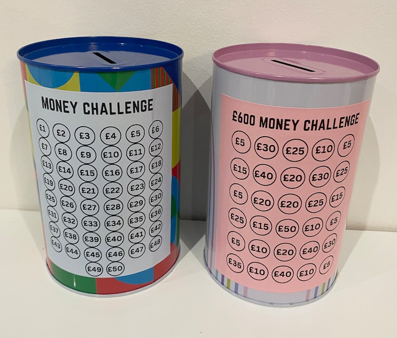 May include: Two metal money challenge piggy banks. The blue and green piggy bank has a money challenge label and a grid of numbers with pound signs. The pink and white piggy bank has a &pound;600 money challenge label and a grid of numbers with pound signs.