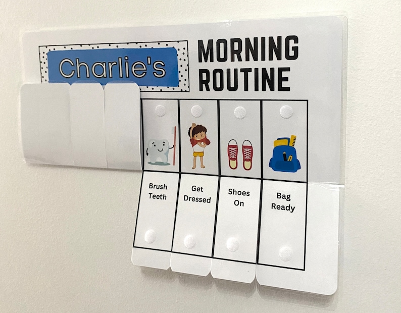 以下が含まれることがあります： 「Charlie's」と「MORNING ROUTINE」の文字が入った白いモーニングルーティンチャート。歯磨き、着替え、靴を履く、バッグの準備のイラストが含まれています。